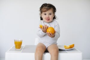 Niña bebé pequeña comiendo cítricos, enseñando sus naranjas con un vaso de zumo de naranja.